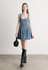 Τζιν φόρεμα με εφαρμοστό κορμό και φαρδιά φούστα, με φαρδιές τιράντες. Φορεμένο με μαύρες μπότες μέχρι το γόνατο και μια μικρή μαύρη τσάντα.