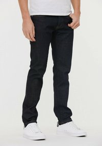 Jeans de mezclilla oscura de corte ajustado, con costuras naranjas y bolsillos frontales, combinados con zapatillas blancas y una camiseta blanca sencilla.