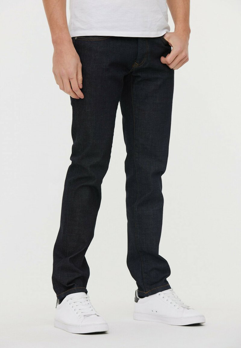 Jeans de mezclilla oscura de corte ajustado, con costuras naranjas y bolsillos frontales, combinados con zapatillas blancas y una camiseta blanca sencilla.