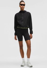 Sort letvægtsjakke med hætte og elastiske manchetter, parret med sorte tætsiddende shorts. Accessoires inkluderer slanke sorte solbriller og hvide sneakers.
