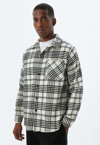 Camisa de franela a cuadros en blanco y negro con botones en el frente y un bolsillo en el pecho; cuenta con un cuello clásico y mangas largas.