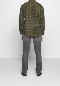 Camisa de manga larga a cuadros verde y rojo con un dobladillo curvado, combinada con jeans grises y zapatillas negras. Tela suave, con los puños abotonados visibles.