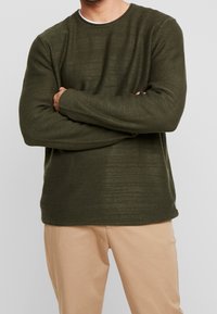 Pull en maille texturée verte à manches longues et col rond, associé à un pantalon beige clair. Détails de coutures visibles à l'ourlet.