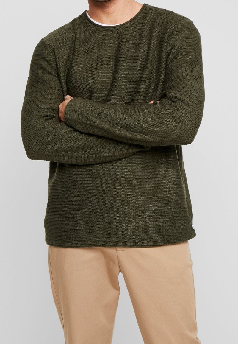 Pull en maille texturée verte à manches longues et col rond, associé à un pantalon beige clair. Détails de coutures visibles à l'ourlet.