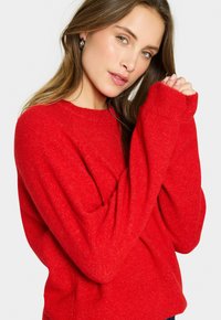 Roter Strickpullover mit lässiger Passform, gerippten Bündchen und Kragen, aus einem strukturierten Stoff, der subtile Tiefe und Wärme verleiht.