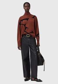 Brązowy sweter z golfem wykonany z dzianiny, z czarnym abstrakcyjnym wzorem, w połączeniu z czarnymi skórzanymi spodniami o szerokich nogawkach i czarnymi płaskimi butami, trzymając czarną torbę.