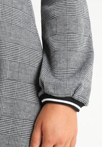 Svartvit mönstrad tyg med ett rutmönster. Kuffad ärm med svartvita randdetaljer vid handleden. Slät textur.