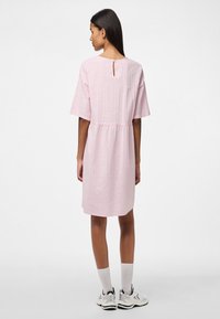 Rozā kleita ar īsām piedurknēm, sakļautu vidukli, strukturētu audumu, atvērumu aizmugurē un līdz ceļiem garu malu. Valkāta ar baltām zeķēm un kedām.