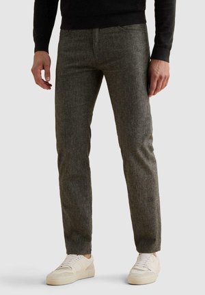 Άνδρας με γκρι παντελόνια slim-fit και λευκά αθλητικά παπούτσια στέκεται μπροστά σε απλό φόντο.