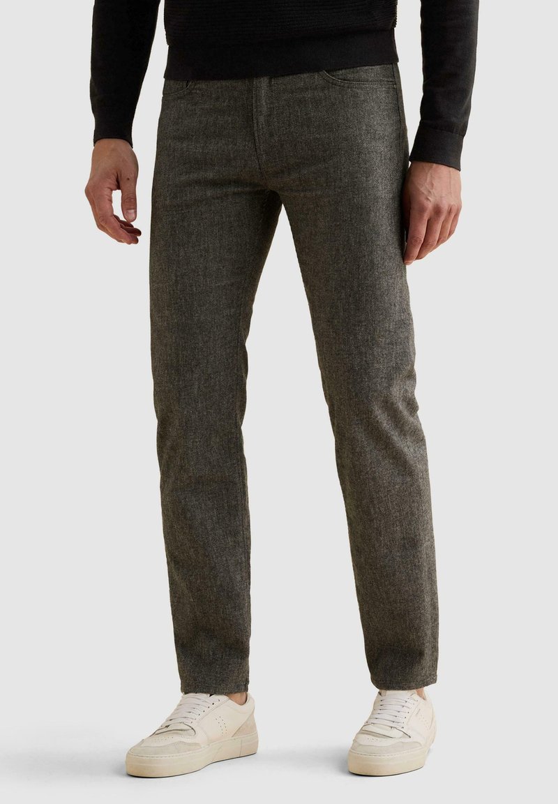 Άνδρας με γκρι παντελόνια slim-fit και λευκά αθλητικά παπούτσια στέκεται μπροστά σε απλό φόντο.