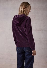 Felpa con cappuccio in corduroy viola, caratterizzata da una texture a coste, con una vestibilità comoda e maniche lunghe, abbinata a jeans in denim blu scuro.