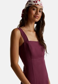 Vestido burdeos sin mangas con escote cuadrado, tirantes planos y textura suave, complementado con un pañuelo de cabeza estampado.