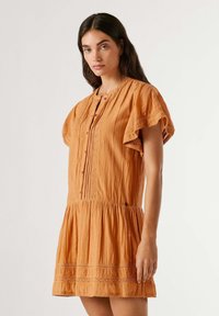 Kvinde med langt mørkt hår iført kortærmet, orange knapklædt kjole med blonde detaljer, stående mod en ensartet lys baggrund.