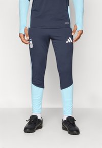 Άνδρας φορά ναυτικά και ανοιχτό μπλε αθλητικά παντελόνια Adidas και μαύρα ποδοσφαιρικά παπούτσια, στέκεται μπροστά σε ένα απλό ανοιχτό φόντο.