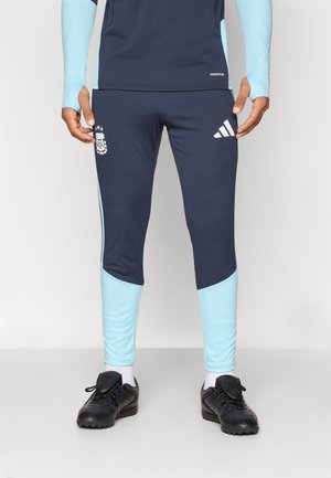 Homme portant un pantalon de sport Adidas bleu marine et bleu clair et des crampons de football noirs, se tenant contre un fond clair et uni.