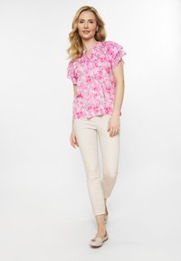 Blomsterfarvet pink bluse med flagrende ærmer, let plissering, parret med beige tætsiddende bukser og lyse slip-on sko.