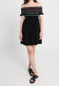 Musta olkaimeton mekko, jossa on smockattu selkäosa ja moniväriset vaakaraitat. Väljä hame ja röyhelöiset hihat. Mustat sandaalit.