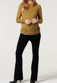 Těhotenské tričko s dlouhým rukávem v hořčicové barvě se staženými boky, na sobě má černé rozšířené kalhoty. Hladká textura, přiléhavý střih a minimalistický design.
