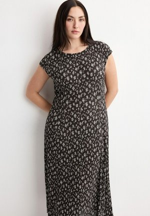 Ženska z dolgimi temnimi lasmi nosi črno obleko z majhnim belim cvetličnim vzorcem in stoji z rokami za hrbtom ob beli steni.