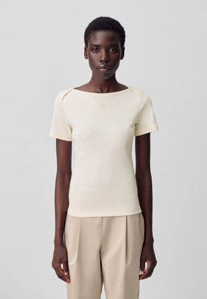 Camiseta ajustada de un beige claro con escote cuadrado, mangas cortas y costuras de hombro sutiles. Presenta un pequeño logo bordado en el pecho.
