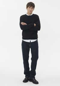 Juodas megztinis, balta palaidinė po juo, tamsiai mėlynos džinsai ir juodi raišteliai. Modelis su sukryžiuotomis rankomis, neutralus fonas.