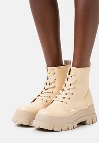 Beżowe za kostkę sznurowane botki wykonane z gładkiej skóry, z masywną, teksturowaną gumową podeszwą i małym akcentem logo po boku.