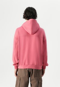 Hoodie rose avec un design spacieux, un capuchon à cordon de serrage et des poignets côtelés. Porté par-dessus un pantalon cargo kaki, mettant en valeur une coupe décontractée et une texture douce.