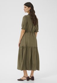 Olivgrön, knälång klänning i mjukt tyg; har puffärmar, elastisk midja och lager på lager-skirt. Bärs med mörkbruna klackskor.