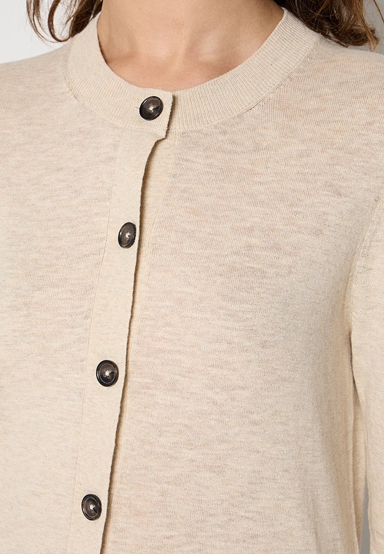 Beżowy kardigan z okrągłym dekoltem, posiadający pięć ciemnych guzików i strukturalną dzianinę. Materiał wydaje się lekki i miękki.