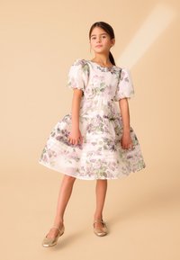 Bela cvetna obleka s vijoličnimi in zelenimi poudarki, nabranimi rokavi in plastmi volanov; nošena z zlatimi baletnimi copatki in postavljena na bež ozadje.