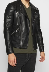 Schwarze Leder-Motorradjacke mit gesteppten Schultern, silbernen Reißverschlüssen und taillierter Passform über einem grünen V-Ausschnitt-Shirt.