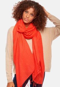 Écharpe rouge en matériau doux, avec un bord frangé et un grand design drapé. Portée de manière décontractée avec un pull beige.