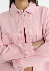 Chaqueta de mezclilla rosa con cuello, botones a presión plateados y dos bolsillos frontales. La tela tiene un patrón texturizado visible.
