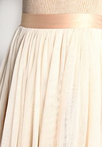 Tylová sukně v jemné krémové barvě s béžovým saténovým pasem. Disponuje plisé texturou, lehkým materiálem a vrstveným designem.