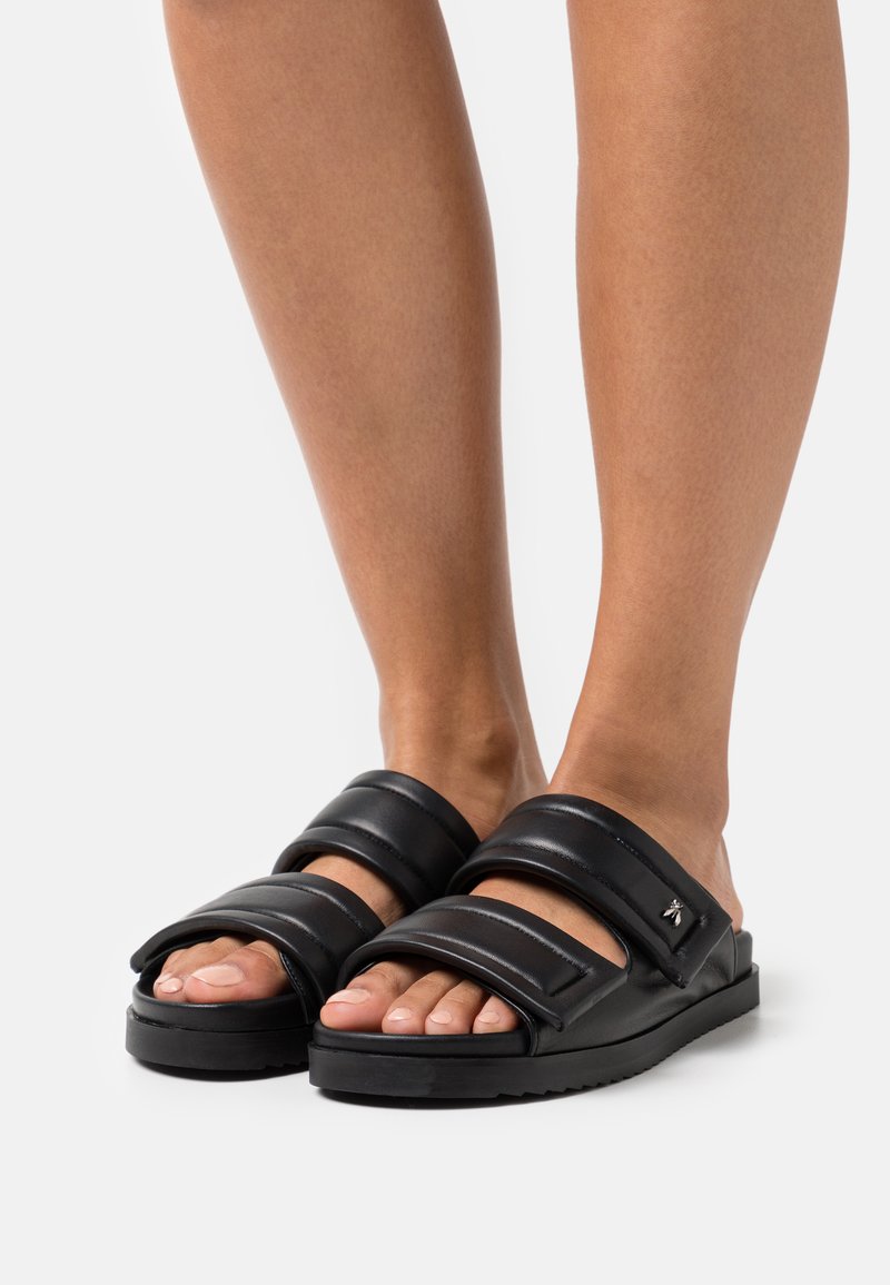 Sorte slide sandaler med to brede stropper, glat lædertekstur, flad sål, sølvhardware accent på siden, der viser bare fødder.