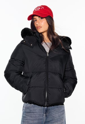 Une femme porte une doudoune noire matelassée à capuche bordée de fourrure, une casquette de baseball rouge et un jean bleu, sur fond blanc.