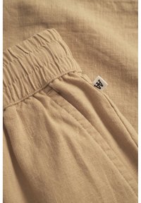 Tessuto di lino beige con vita elasticizzata arricciata e dettagli plissettati. Presenta un piccolo etichetta con il marchio sul lato. Aspetto morbido e testurizzato.