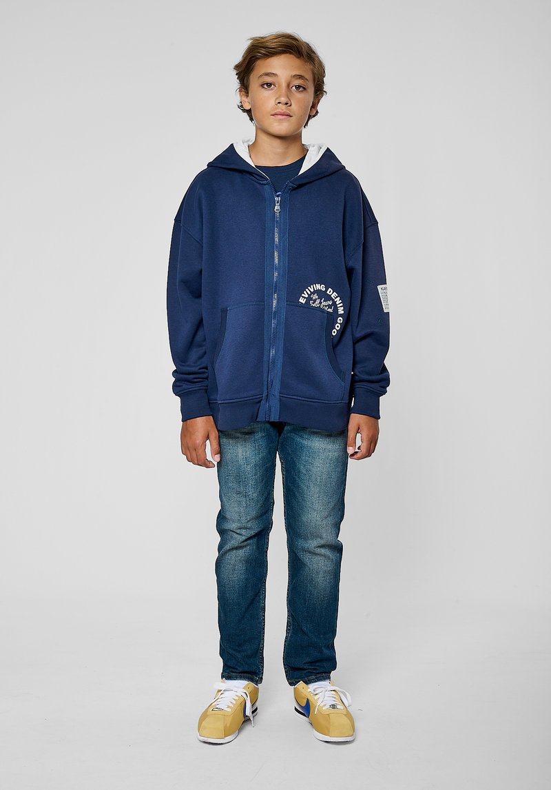Sweat à capuche navy zippé avec doublure grise, poches avant et détail de logo ; associé à un jean bleu délavé et des baskets jaunes.