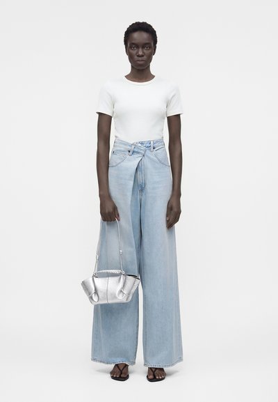 Γυναίκα με λευκή μπλούζα με κοντά μανίκια, ανοιχτό μπλε τζιν παντελόνι με φαρδιές γραμμές και ψηλή μέση, μαύρα σανδάλια, κρατώντας μικρή ασημένια τσάντα, στέκεται μπροστά σε απλό φόντο.