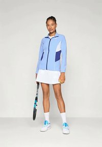 LyseblåIdrettsjakke med hvite og marineblå detaljer, parret med en hvit skjørt. Modellen holder en tennisracket og har på seg joggesko med blå detaljer.