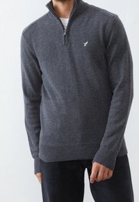 Grauer Pullover mit Reißverschluss, gerippten Bündchen und Saum, mit einem kleinen gestickten Logo auf der Brust. Hergestellt aus einem strukturierten Strickstoff.