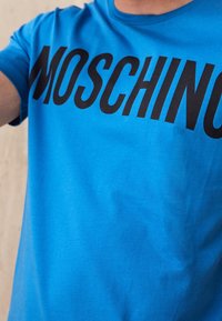 Modré tričko s velkým černým textem "MOSCHINO" přes hruď, které nosí osoba na béžovém pozadí.