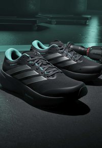 Paire de chaussures de course Adidas noires avec doublure intérieure bleu clair sur le sol de la salle de sport, à côté d'un rouleau de fitness noir et rouge.