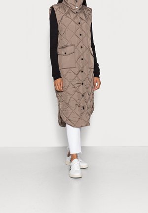 Sleeveless long quilted taupe vest with a high collar and black buttons over black long sleeves, white cropped pants, and white sneakers.