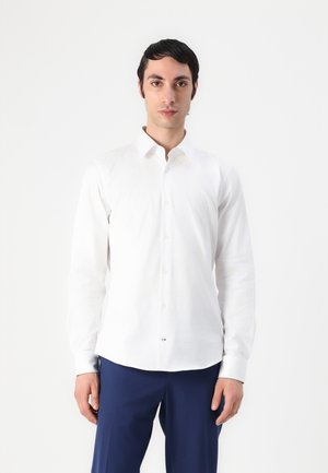 Chemise blanche à boutons, en tissu lisse, avec un col classique, des manches longues et un ourlet incurvé. Portée avec un pantalon navy.