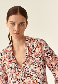 Blomstret blouse med krave, der har livlige tryk i pink, blå, rød og marineblå på et let stof. Knapdetalje synlig.