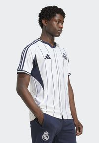 Jersey blanco de manga corta con rayas verticales en navy, que presenta un logo de Adidas y el escudo del Real Madrid en el pecho, combinado con pantalones navy.