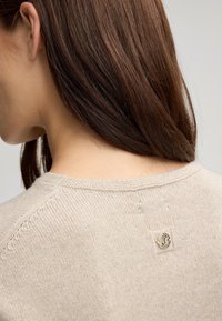 Maglione beige in maglia con una texture liscia e un colletto rotondo. Presenta una piccola toppa logata in rilievo sul retro vicino al colletto.