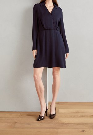 Knee-langer, navyblauer Wickelkleid mit langen Ärmeln und Kragen, das eine taillierte Taille und einen ausgestellten Rock hat. Kombiniert mit braunen Pumps mit spitzem Fuß.
