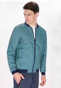 Mannen bomberjack in teal met een geometrisch patroon. Geribbelde donkerblauwe kraag en manchetten, ritssluiting en zijzakken voor functionaliteit.